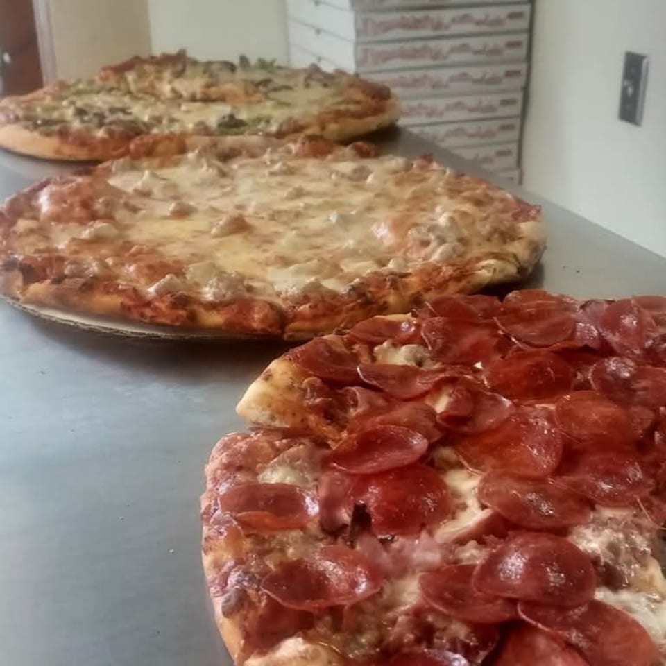 Three Roma Pizza pizzas on a metal counter, with pepperoni, sausage, and mushroom toppings.